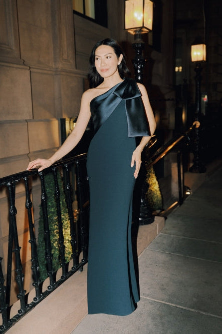 A woman wearing a floor-length dark teal one-shoulder gown with a large satin bow detail on the shoulder, standing on a city sidewalk at night next to a black metal railing.