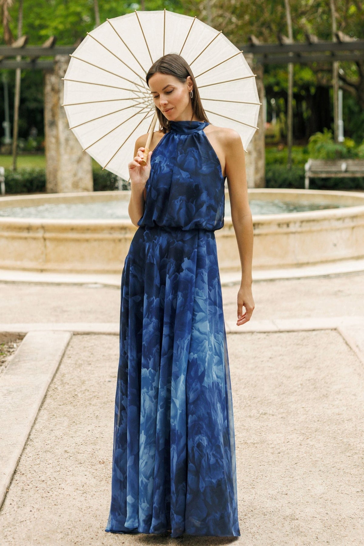 Woman wearing the Tami Chiffon floral printed halter neck evening gown, holding a parasol and posing in front of a fountain. 